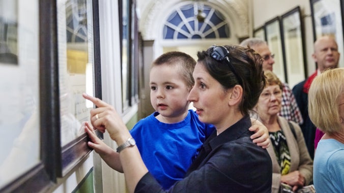 Mam yn dal ei phlentyn ac yn pwyntio at arddangosiad o luniau ar waliau cyntedd yn y tŷ yn Erddig, Clwyd.